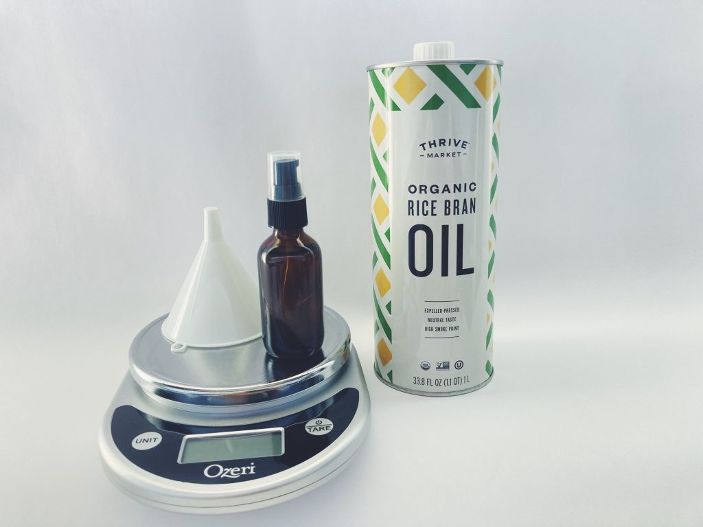 Small bottle of DIY cleansing oil and a funnel sit on a kitchen scale next to a large bottle of organic rice bran oil on a white background.