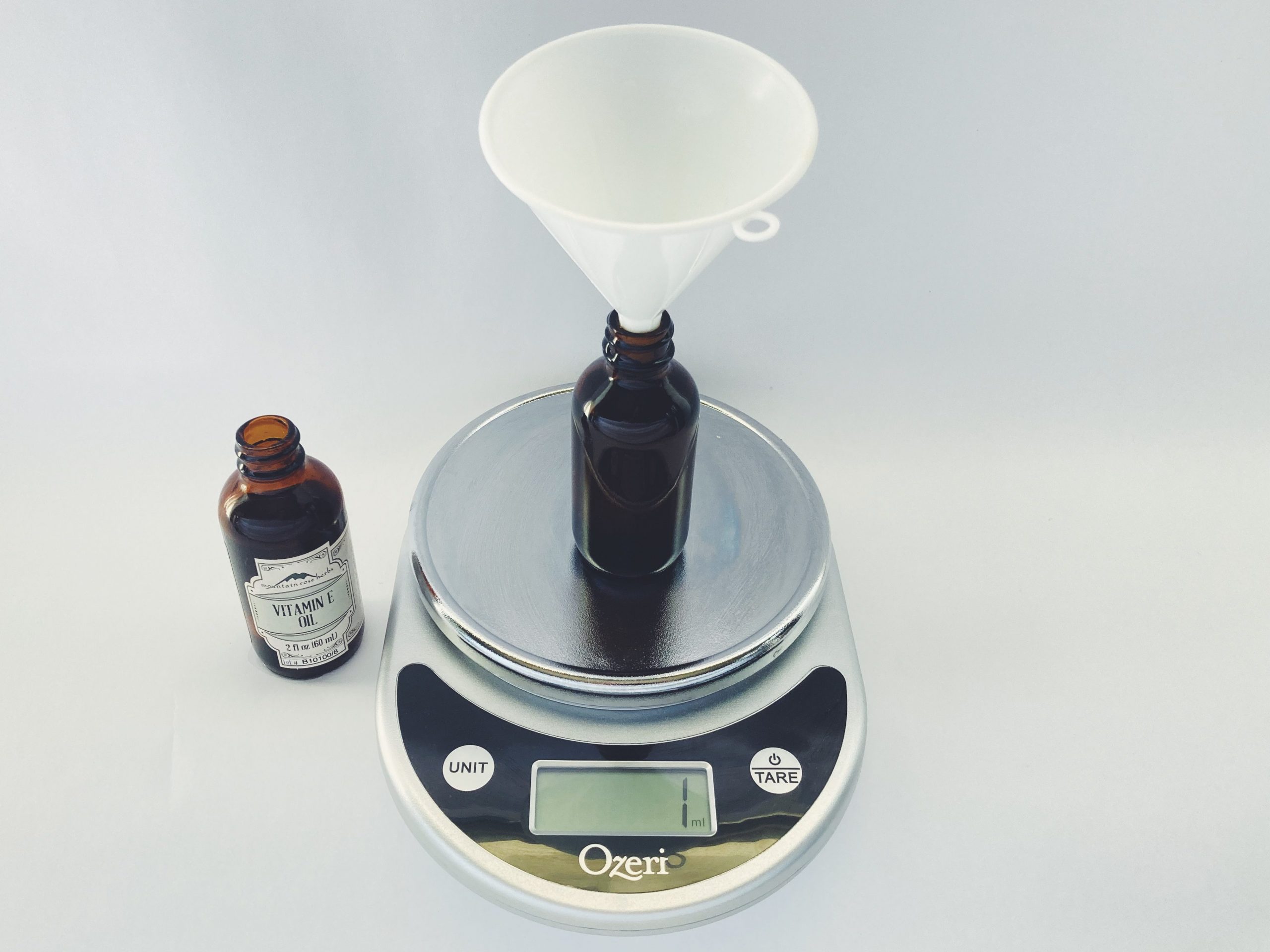A funnel sitting in a glass jar for DIY cleansing oil on a kitchen scale. A glass bottle of Vitamin E oil sits next to the kitchen scale.