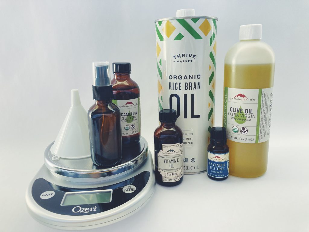 Supplies for DIY Cleansing Oil including a kitchen scale, funnel, glass bottle, camellia oil, vitamin e oil, organic rice bran oil, lavender tea tree essential oil, and olive oil on a white background.