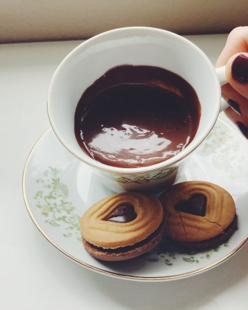 Teacup of drinking chocolate with two cookies next to it being tilted to sip.